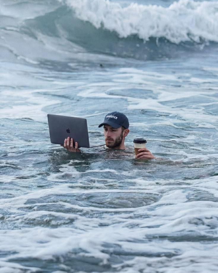 Someone working on a laptop while standing in the sea, holding a coffee